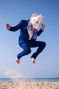 A man in a suit wearing a unicorn mask is jumping on the beach